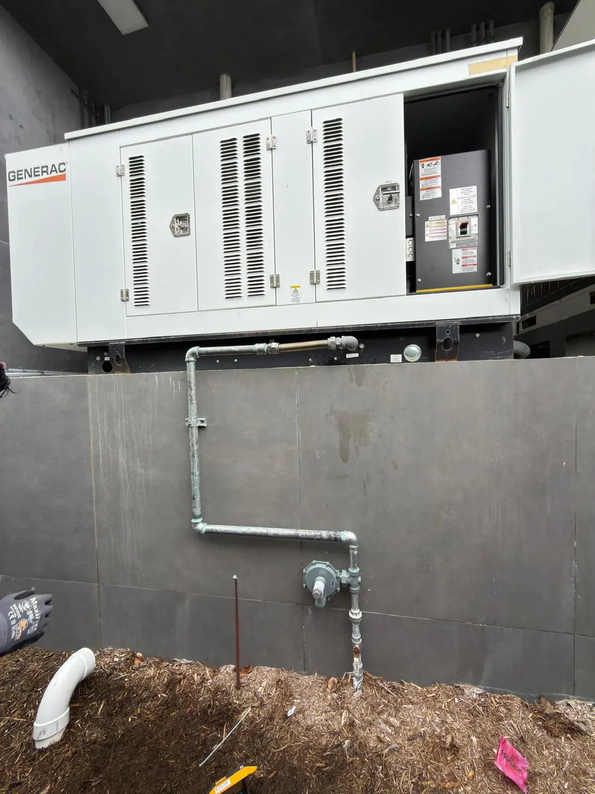 Large white enclosed generator on a raised platform with a galvanized fuel line and regulator on the wall below