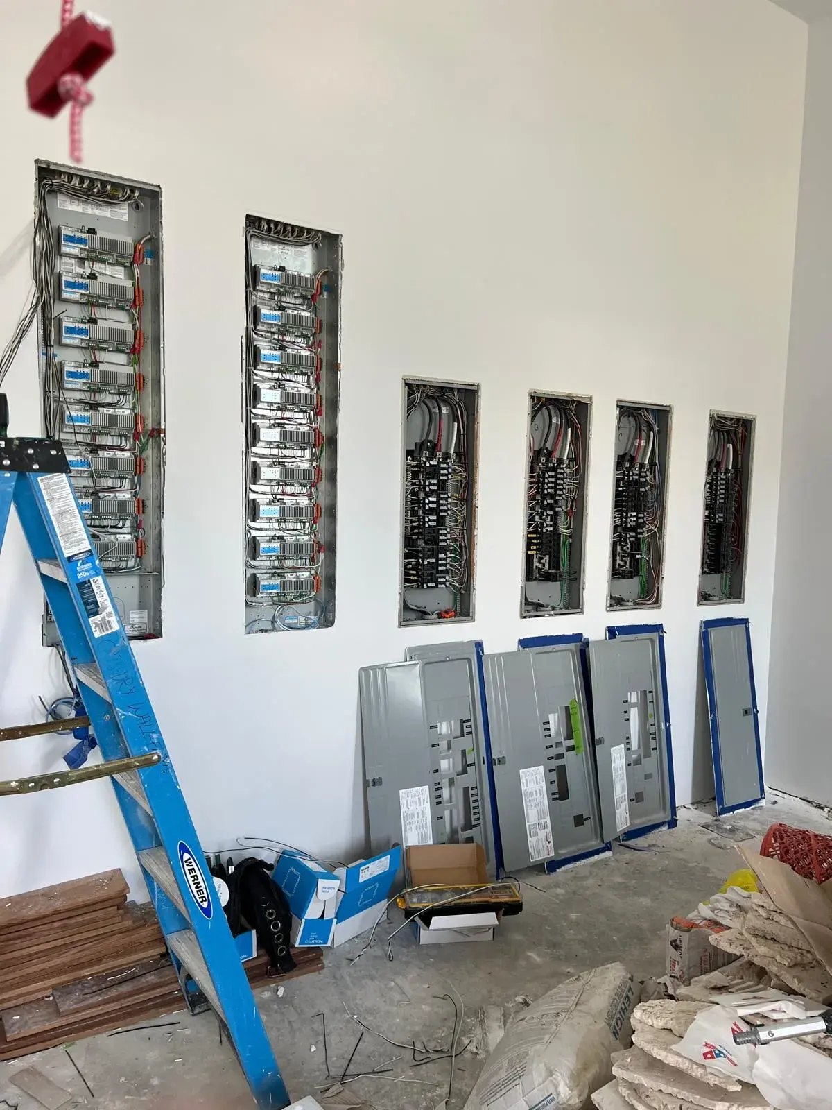 White wall with two tall lighting control enclosures and four open breaker panels recessed in it, covers leaning below