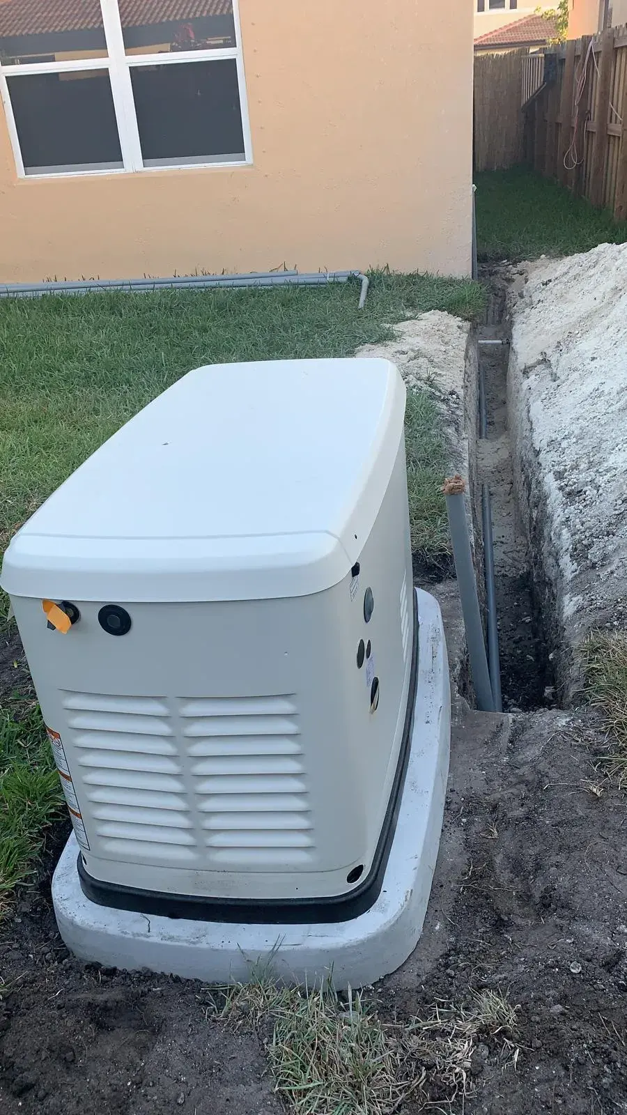 Standby generator on a concrete pad beside a house, with the conduit trench being prepared