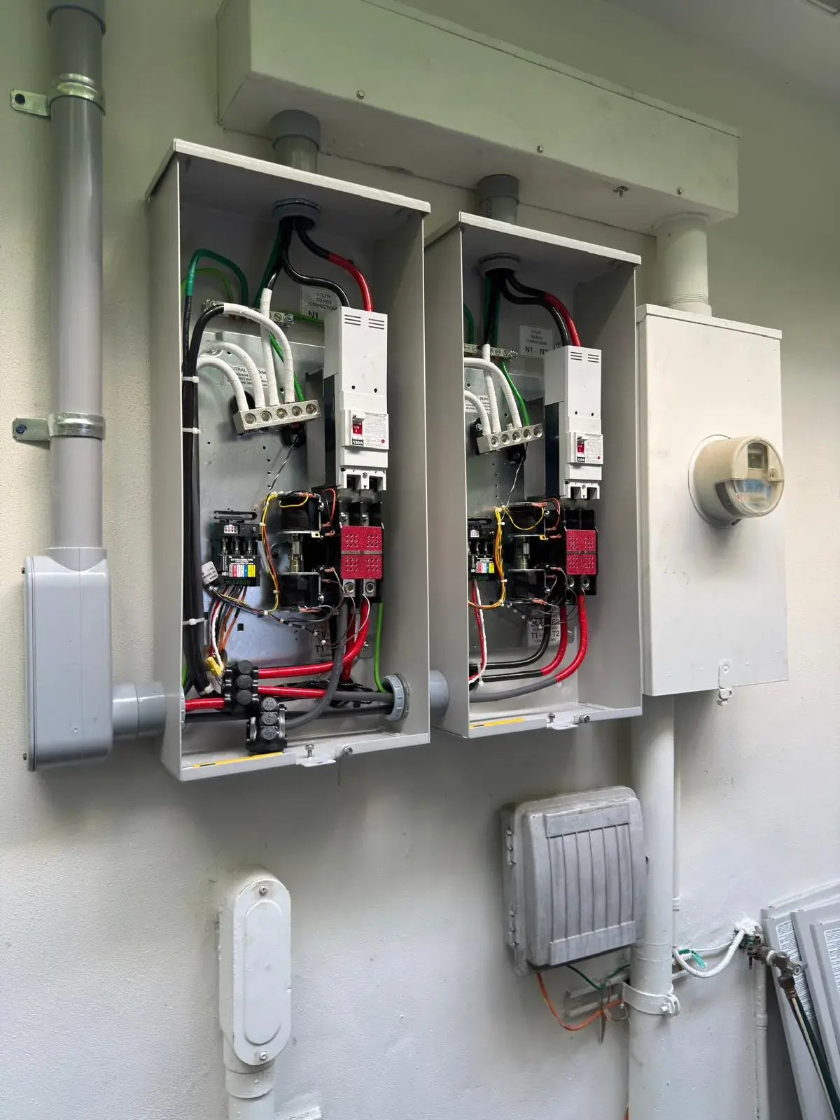 Two open transfer switch enclosures side by side next to a utility meter on a white wall