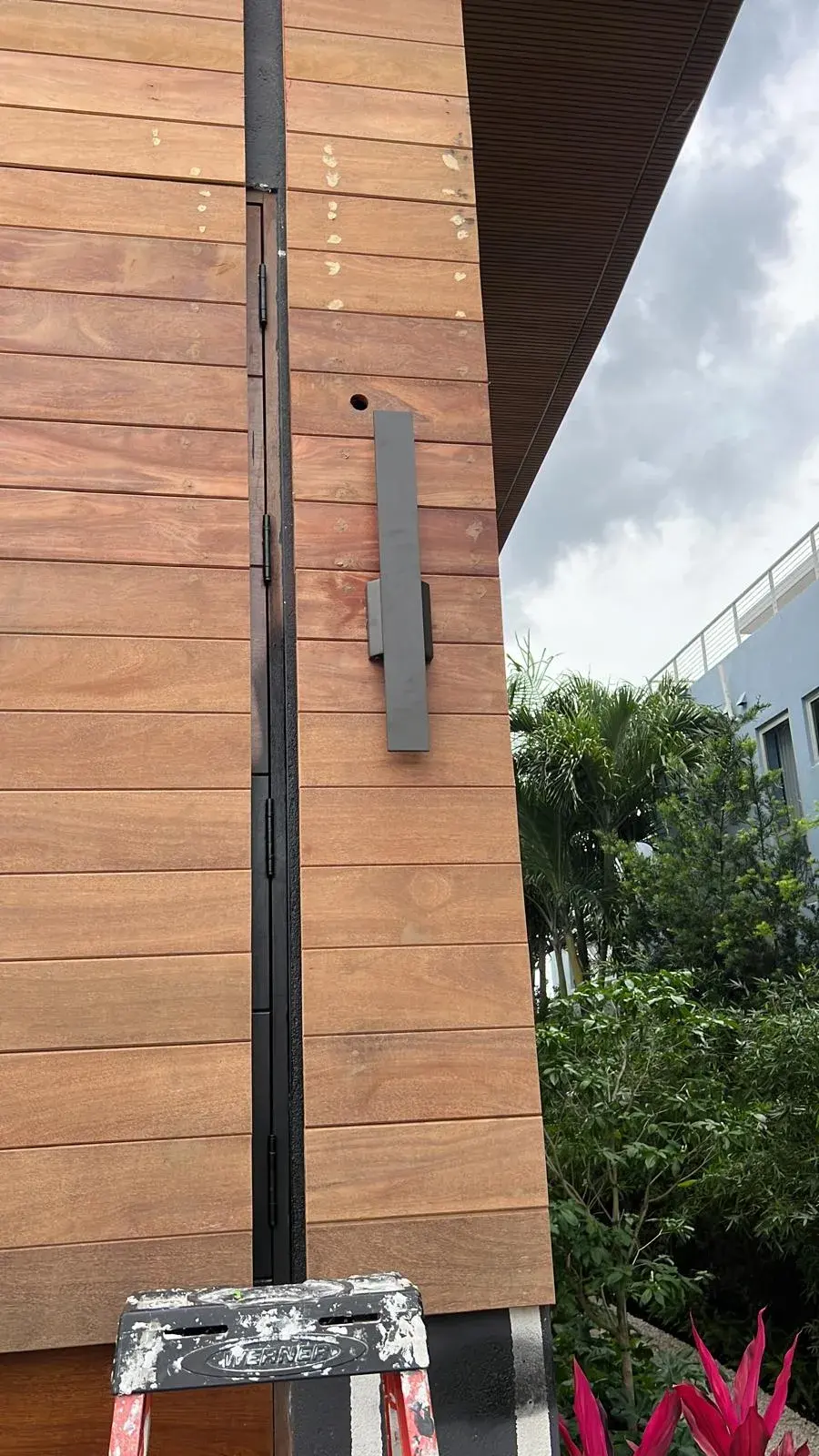 Black linear wall sconce on horizontal wood siding, with a ladder below and tropical plants to the side