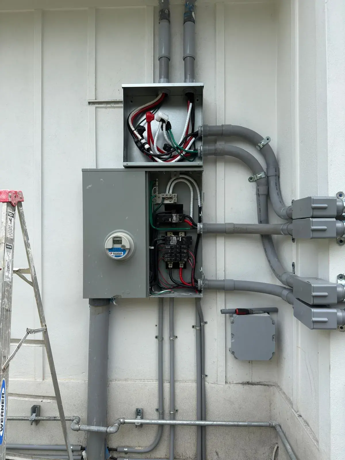 Open meter and main panel enclosure below a pull box, with gray conduit runs on a white wall